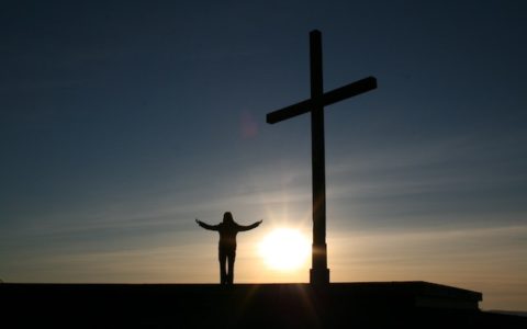 Silhouette of a person standing with arms stretched out in front of a cross at sunset